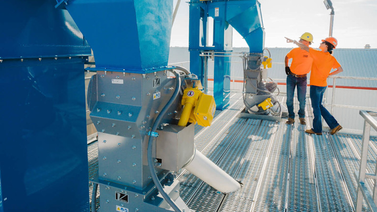 Two workers in orange shirts beside a machine, having a conversation and overseeing its operation