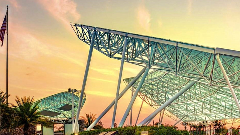 Modern glass and steel canopy structure with angled supports against a golden sunset sky, American flag visible, landscaped garden path below